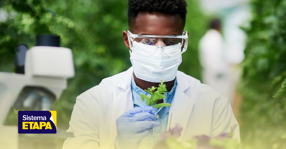 Estudante participando de uma atividade científica relacionada à Biologia na Olimpíada Internacional de Biologia (IBO), usando máscara de proteção, óculos de segurança e luvas em um ambiente com plantas ao fundo.