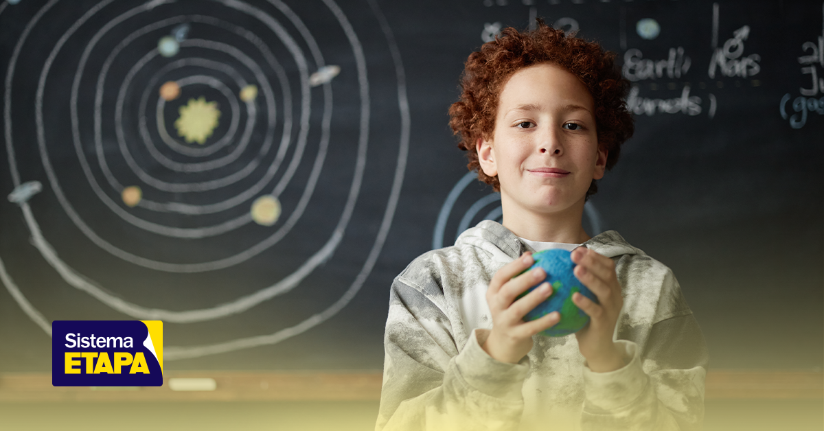 Jovem estudante participando da Olimpíada Internacional de Astronomia e Astrofísica (IOAA), segurando um globo terrestre com fundo de quadro negro com desenhos do sistema solar.